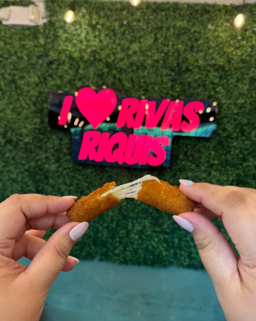 Dos manos parten un mozzarella stick frente al rótulo I love Rivas RiQUi's